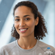 Headshot of a smiling, diverse woman in her late 20s or early 30s, looking confidently at the viewer with a soft blue backgro